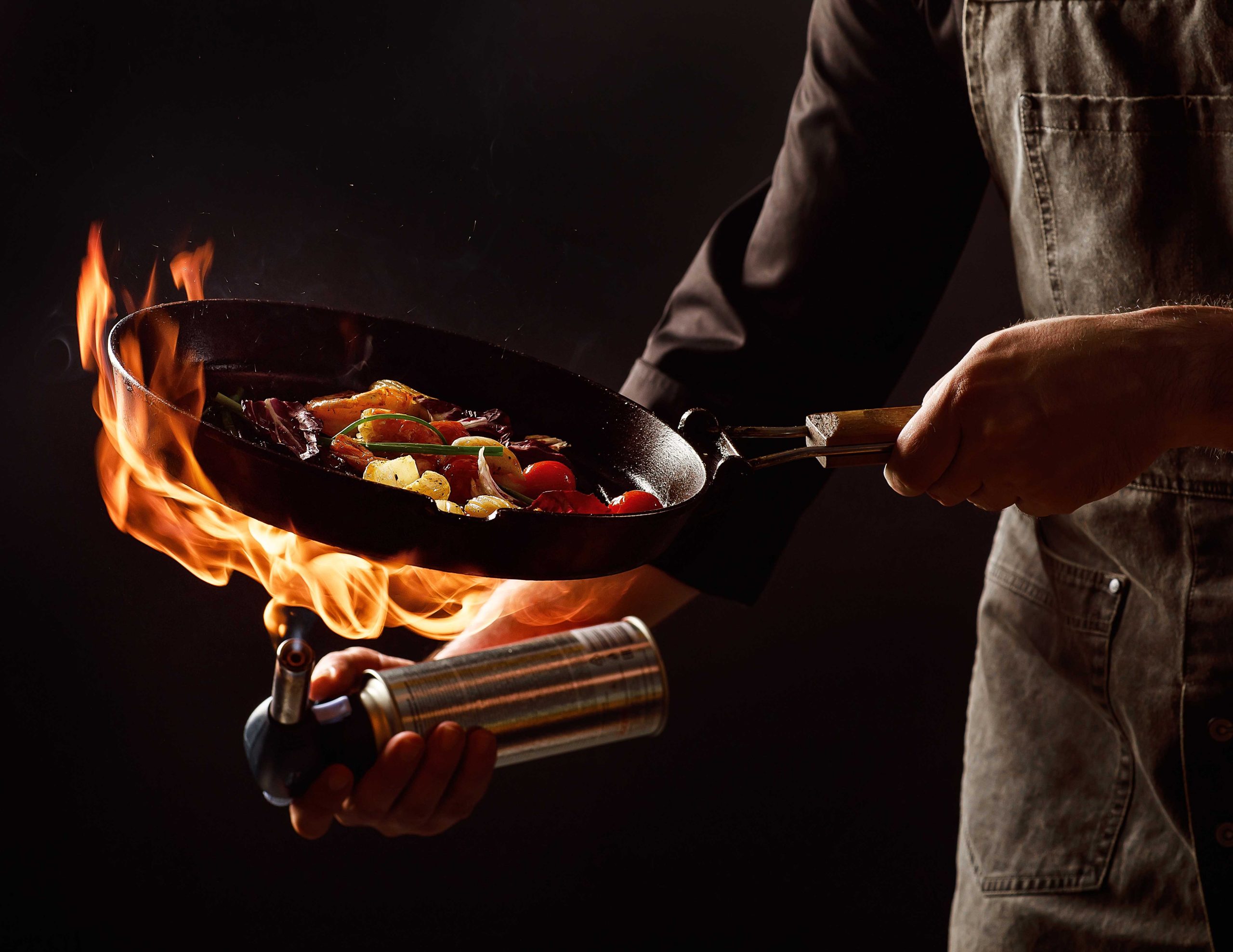 Chef cooks with fire in a pan shrimp with vegetables. On a black background, restaurant service